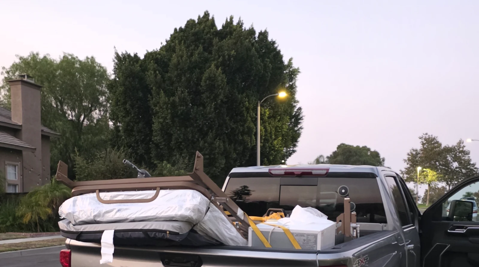 A silver Z Junk 4x4 pickup with a bed rack loaded with a wrapped mattress and boxes for a bulky-item pickup on a residential street at dusk.